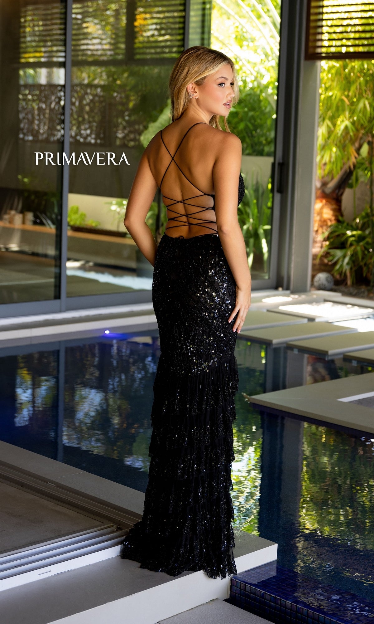 A woman in the Primavera Long Beaded Ruffled Prom Dress 4122 stands near an indoor pool, with